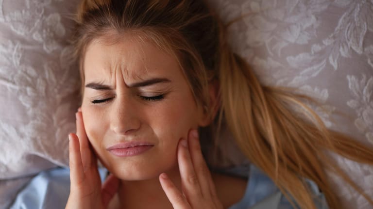 Junge Frau liegt im Bett mit Kieferschmerzen: Beim Zähneknirschen reiben oder pressen Betroffene ihre Zähne aneinander, das schadet auf Dauer dem Zahnschmelz und Zahnfleisch.