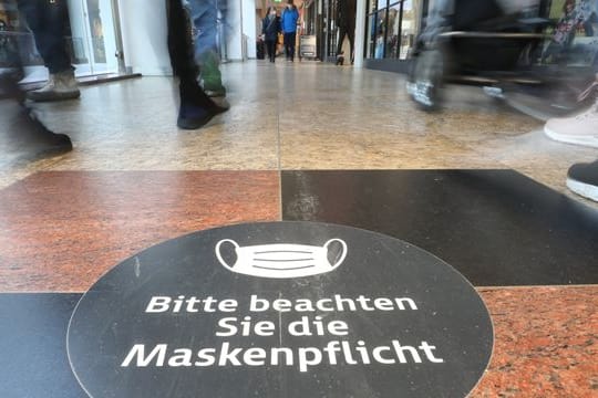 Passanten laufen durch ein Einkaufszentrum auf dessen Fußboden ein Hinweisschild zur Maskenpflicht aufgeklebt ist.