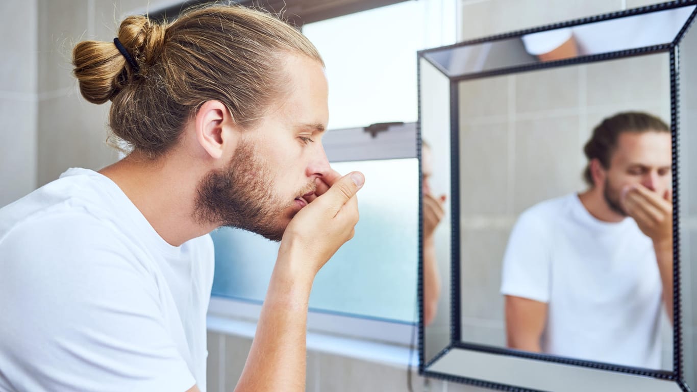Mann vor Spiegel testet ob er Mundgeruch hat: Wenn eine gute Mundhygiene gegen Mundgeruch nicht ausreicht und Begleitsymptome erscheinen, sollte ein Arzt die Ursache klären.