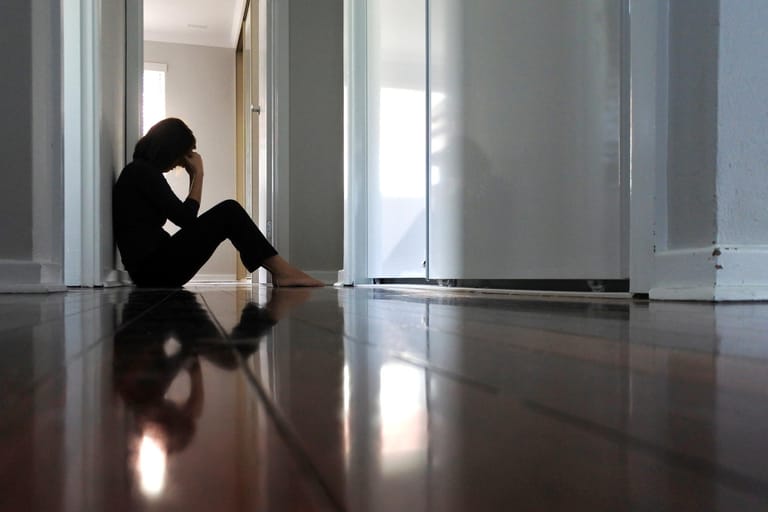 Depressive Frau sitzt auf dem Boden im Flur: Seelisches Leiden: Stress, Nervosität und andere psychische Leiden können ebenfalls eine Ursache für Scheidentrockenheit sein. Sexuelle Erregung sorgt normalerweise für die Befeuchtung der Scheide beim Geschlechtsverkehr. Diese lässt jedoch bei seelischen Problemen nach.