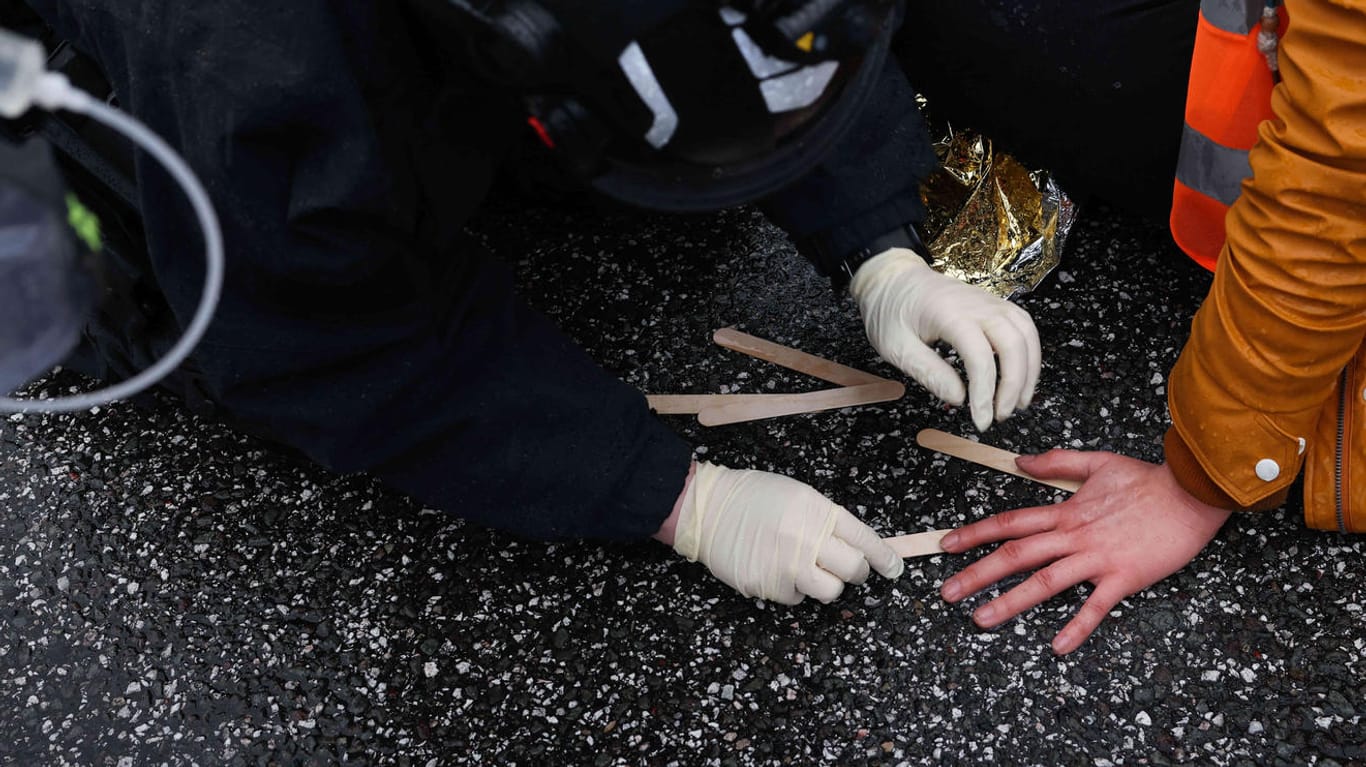 Eine Beamtin des technischen Zuges der Polizei löst mit einer Chemikalie und Holzspachteln die festgeklebte Hand einer Klimaaktivistin von der Fahrbahn.