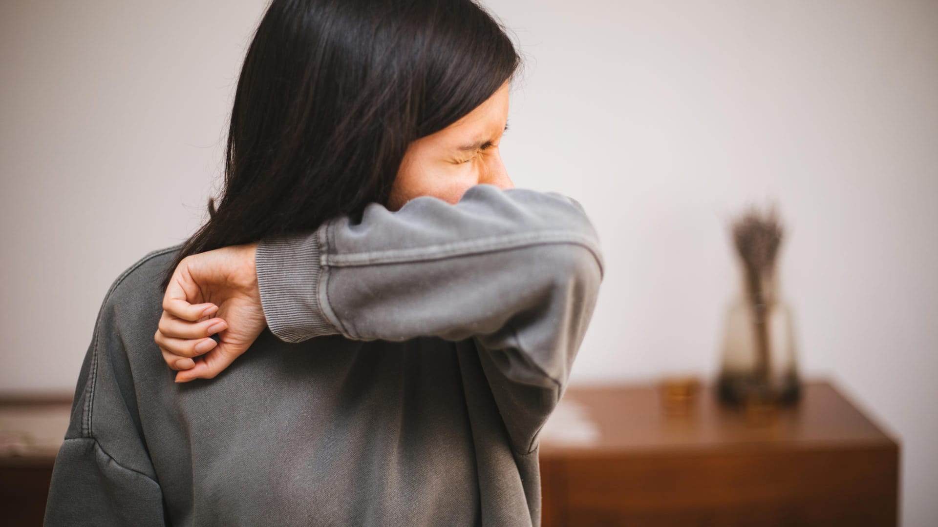 Junge Frau niest in ihren Ellenbogen: Belastungsinkontinenz: Bei dieser Form kommt es bei Belastung zu plötzlichem Urinverlust. Betroffene können beim Niesen oder Treppensteigen ihren Harndrang nicht mehr kontrollieren. Ursache ist meist eine Schwäche der Beckenbodenmuskulatur.