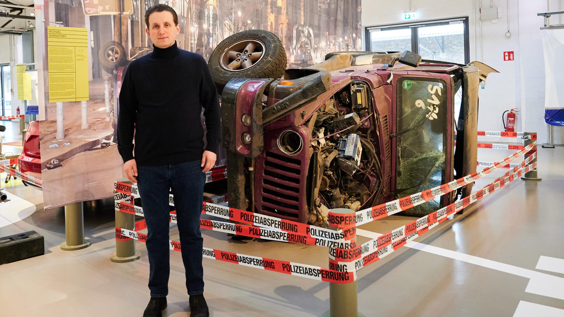 M. Warshitskys Sohn Maximilian steht vor dem Jeep seines Vaters, in welchem dieser als Opfer der Kudamm-Raser 2016 ums Leben kam: Im Hintergrund des Wracks ist der echte Tatort zu sehen.