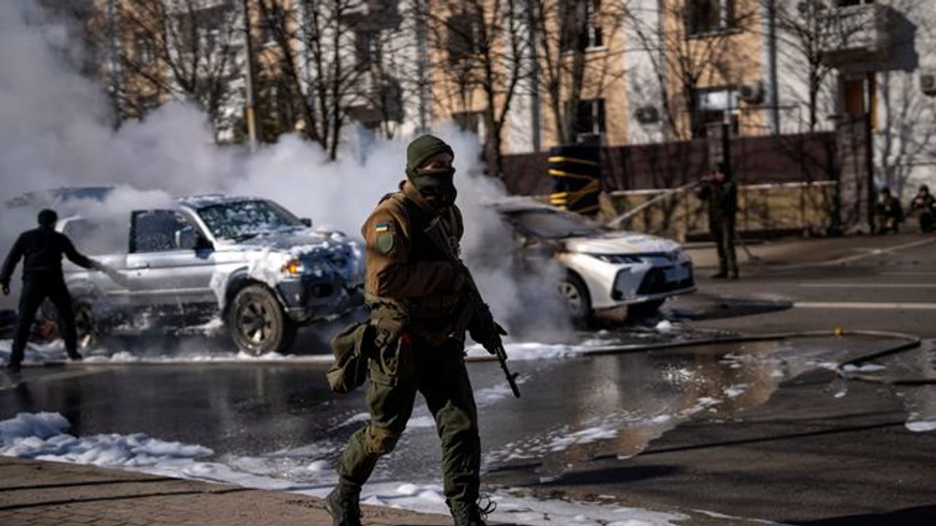 Ein ukrainischer Soldat bezieht Stellung vor einer Militäreinrichtung, während Feuerwehrleute im Hintergrund zwei brennende Autos löschen.