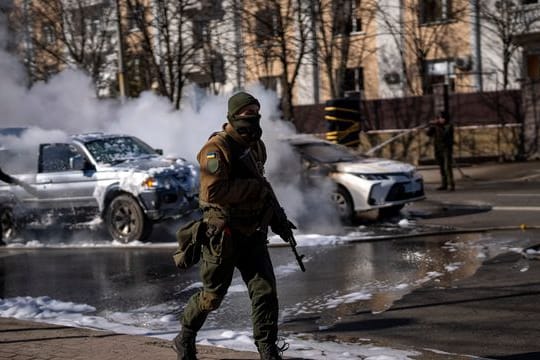 Ein ukrainischer Soldat bezieht Stellung vor einer Militäreinrichtung, während Feuerwehrleute im Hintergrund zwei brennende Autos löschen.