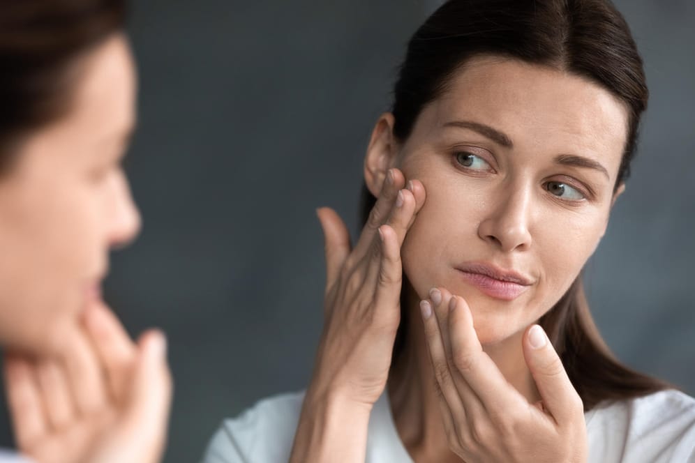 Eine Frau betrachtet ihr Gesicht im Spiegel. Psychische Belastungen können sich negativ auf das Hautbild auswirken.