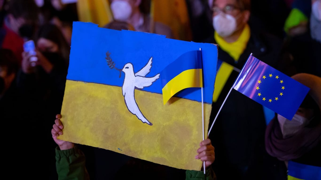 In München hält ein Kind Schilder mit einer Friedenstaube und der Flagge der Ukraine in die Höhe.