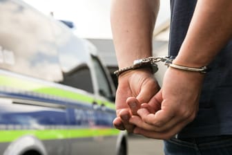 Ein Mann in Handschellen vor einem Polizeiauto (Symbolbild): Die Polizei ermittelt insgesamt gegen neun Verdächtige.
