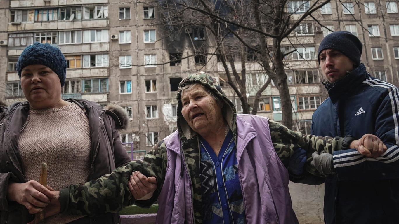 Menschen helfen einer älteren Frau beim Gehen auf einer Straße mit einem von Granaten getroffenen Wohnhaus im Hintergrund in Mariupol