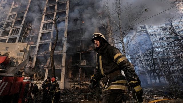 Einsatzkräfte der Feuerwehr stehen in Kiew vor einem brennenden Wohngebäude, dass durch Bombenbeschuss des russischen Militärs zerstört wurde.