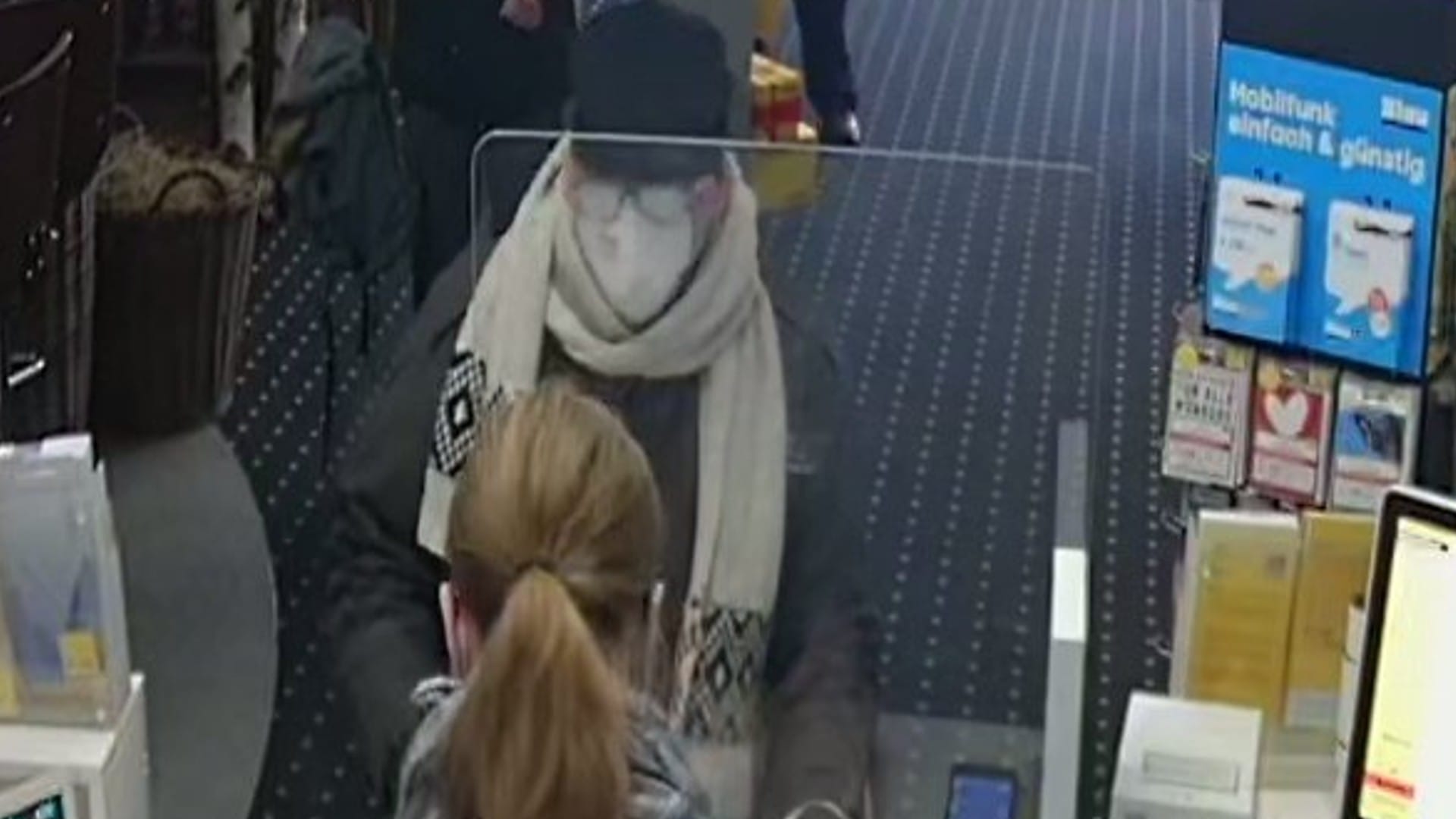 Ein weiteres Bild der Überwachungskamera: Der Mann trug eine Mund-Nasen-Bedeckung, eine Brille, eine Schiebermütze, einen langen dunklen Mantel, eine dunkle Hose, dunkle Schuhe, schwarze Lederhandschuhe und einen auffälligen beigen Schal.