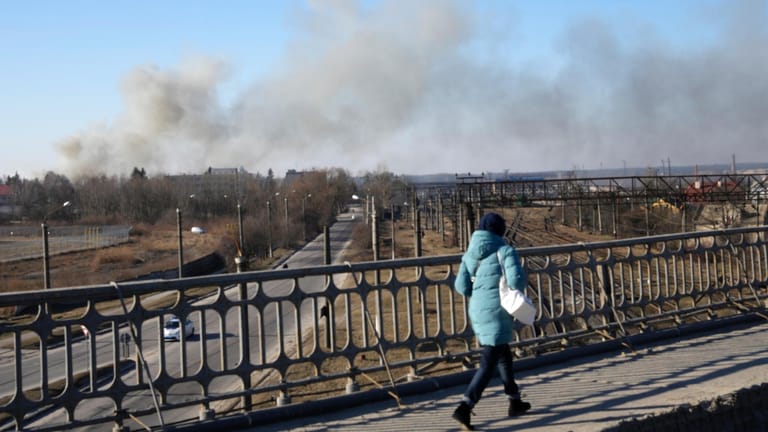 18. März 2022: Eine Frau geht über eine Brücke, während eine Rauchwolke nach einer Explosion aufsteigt. Nach Angaben des Bürgermeisters von Lwiw schlugen russische Raketen in der Nähe des Flughafens der Stadt ein.