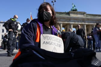 Eine Aktivistin der "Letzten Generation" vor dem Brandenburger Tor mit einem Schild um den Hals, das darauf hinweist, dass sie sich festgeklebt hat: Als Grund für die Proteste am Freitag gilt die erreichte 100-Tage-Marke der Bundesregierung.
