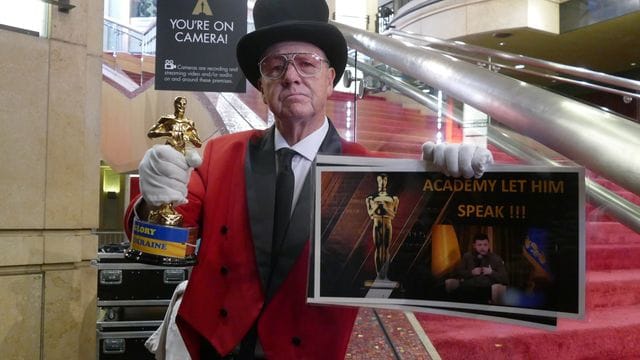 Touristenführer Gregg Donovan hält ein Schild mit der Aufschrift "Academy Let Him Speak" mit einem Bild vom ukrainischen Präsidenten Selenskyj.