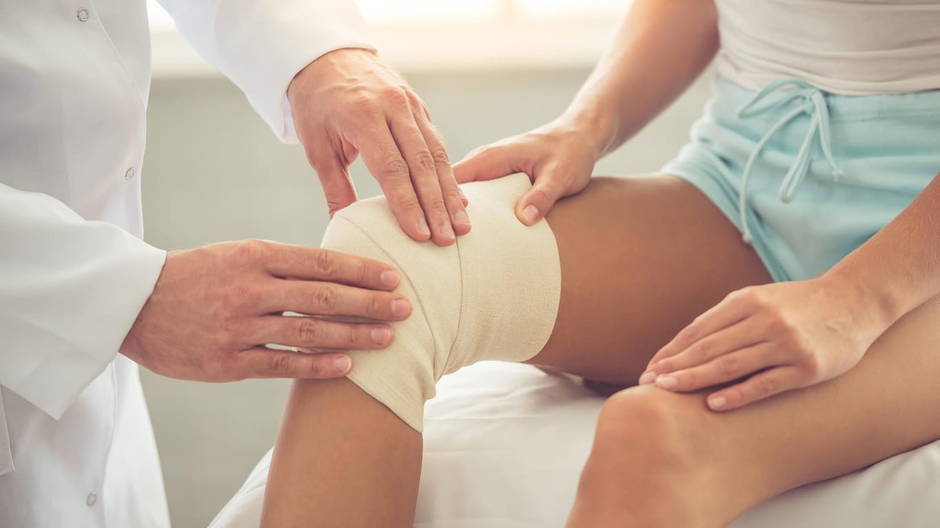 Art untersucht verletztes Knie einer Frau: Nach einer Operation dauert die Wundheilung oft länger. Besonders größere Verletzungen benötigen eine professionelle Wundpflege und viel Geduld.