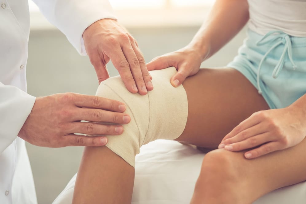 Art untersucht verletztes Knie einer Frau: Nach einer Operation dauert die Wundheilung oft länger. Besonders größere Verletzungen benötigen eine professionelle Wundpflege und viel Geduld.