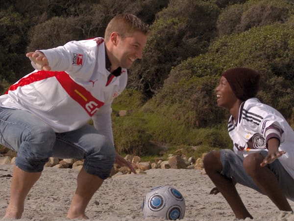 Thomas Hitzlsperger und ein afrikanischer Junge spielen am Strand Fußball.