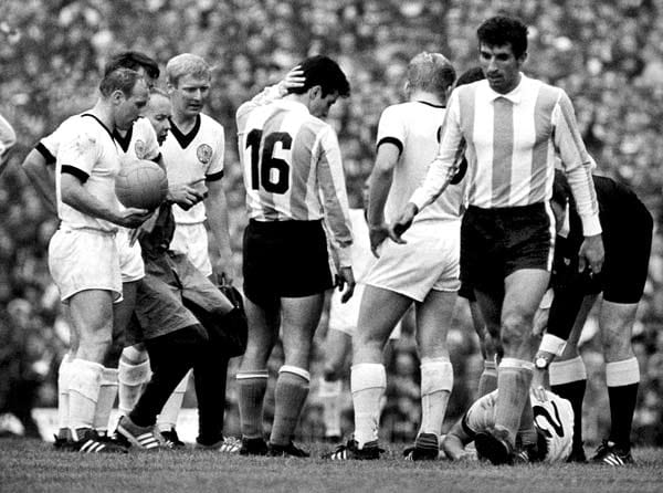 Gänzlich kalt lässt es den Argentinier Alberto González (Nummer 16) am 16. Juli 1966 in Birmingham nicht, dass sich sein deutscher Gegenspieler Horst-Dieter Höttges (Nummer 2) vor Schmerzen am Boden krümmt. Die DFB-Stars Uwe Seeler, Wolfgang Overath, Karl-Heinz Schnellinger und Helmut Haller (alle in Weiß, von links) sorgen sich ebenfalls um ihren Verteidiger.