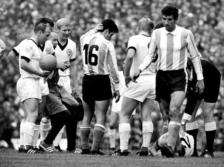 Gänzlich kalt lässt es den Argentinier Alberto González (Nummer 16) am 16. Juli 1966 in Birmingham nicht, dass sich sein deutscher Gegenspieler Horst-Dieter Höttges (Nummer 2) vor Schmerzen am Boden krümmt. Die DFB-Stars Uwe Seeler, Wolfgang Overath, Karl-Heinz Schnellinger und Helmut Haller (alle in Weiß, von links) sorgen sich ebenfalls um ihren Verteidiger.