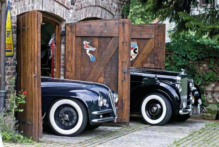 Hier ist eine alte Garge mit Backsteinmauern zu sehen. Vorne ragt ein Alfa Romeo 6C 2500SS Coupé und dahinter ein Bentley Mark VI mit Sonderkarosserie hervor.
