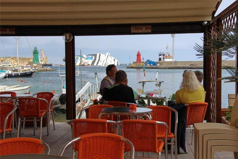 Gäste in einem Hafenrestaurant von Giglio mit Blick auf das havarierte Kreuzfahrtschiff. Der Bürgermeister hat immerhin Schilder anbringen lassen, dass die verehrten Gäste doch bitte den Respekt vor den Opfern der Tragödie und ihren Familien nicht vergessen mögen.