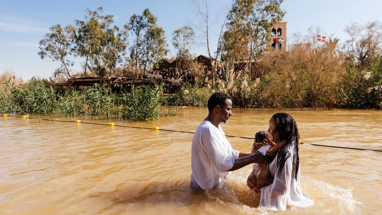 Getauft: Eine Frau mit weißem Gewand taucht mit ihrem Kind im Arm auf dem Fluss auf.