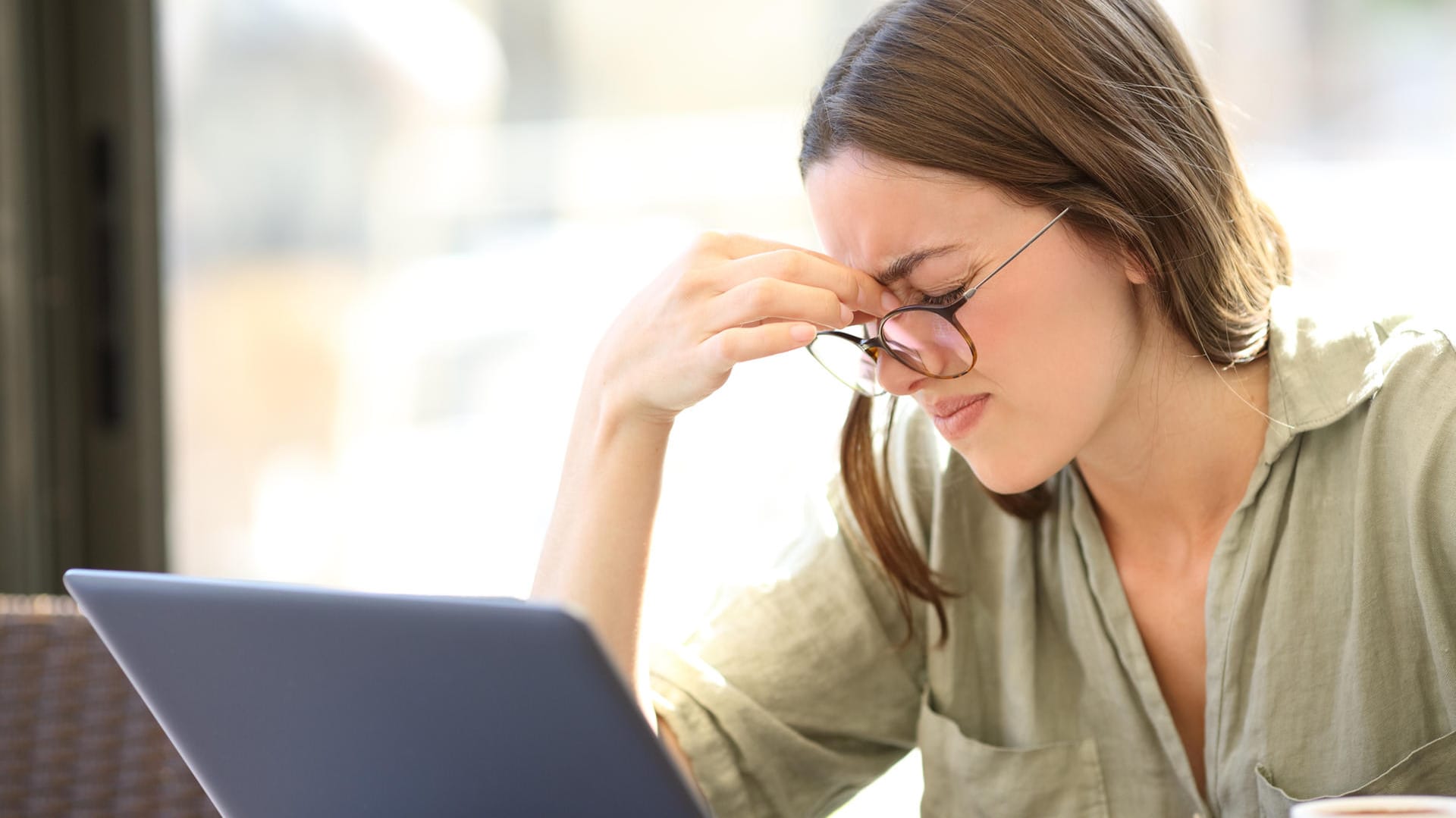 Junge Frau vor Laptop kneift die Augen zu. Im Homeoffice sind die Lichtverhältnisse oft schlechter als im Büro. Darunter leiden die Augen.