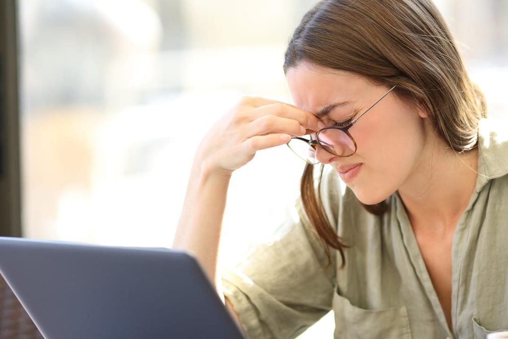 Junge Frau vor Laptop kneift die Augen zu. Im Homeoffice sind die Lichtverhältnisse oft schlechter als im Büro. Darunter leiden die Augen.