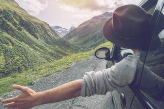 Eine Person fährt mit einem Auto durch die Berge: In einem Mietwagen lassen sich Urlaubsorte bequem erkunden.