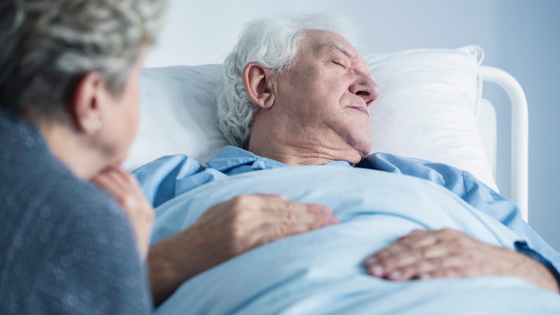 Eine Frau sitzt am Krankenbett ihres Mannes.