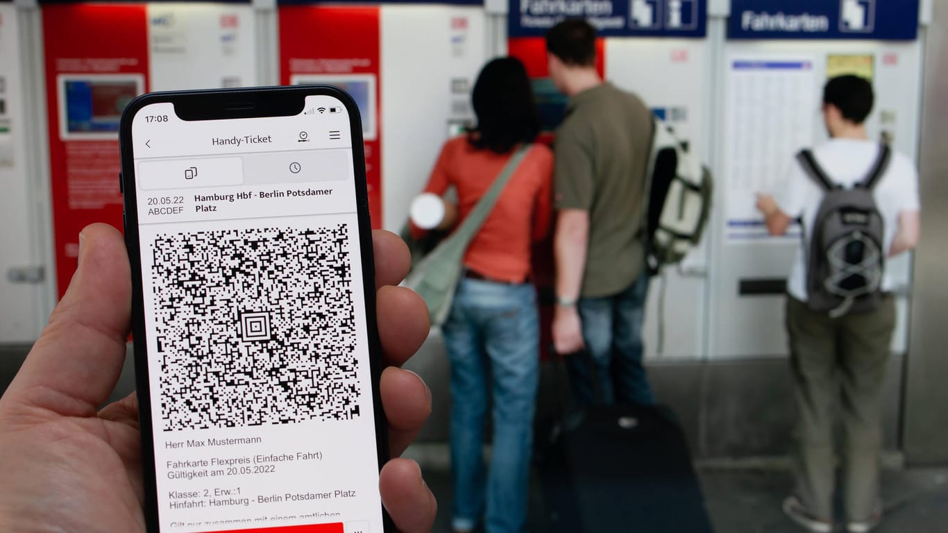 Bahnticket auf einem Mobiltelefon. Im Hintergrund kaufen Personen Tickets an einem Automaten.
