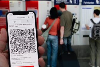Bahnticket auf einem Mobiltelefon. Im Hintergrund kaufen Personen Tickets an einem Automaten.