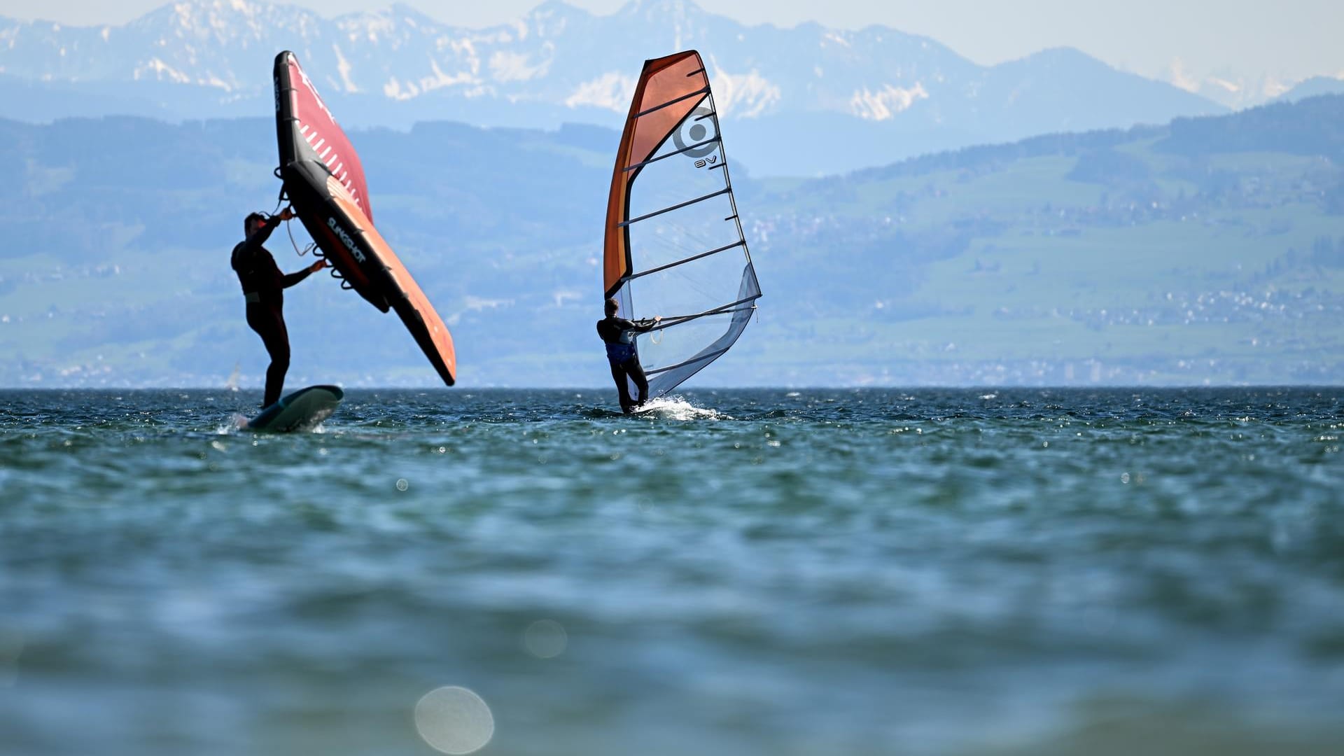 Zwei Surfer gleiten vor Immenstaad über den Bodensee, während im Hintergrund die Alpen in der Schweiz zu sehen sind.