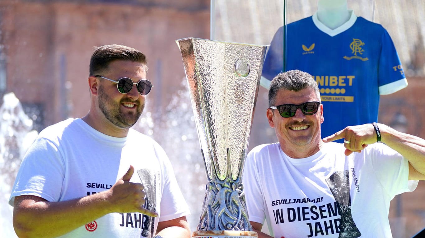 Zwei Frankfurter Fans posieren mit der Trophäe der Europa League; im Hintergrund wacht das Trikot des Finalgegners Glasgow Rangers über sie.