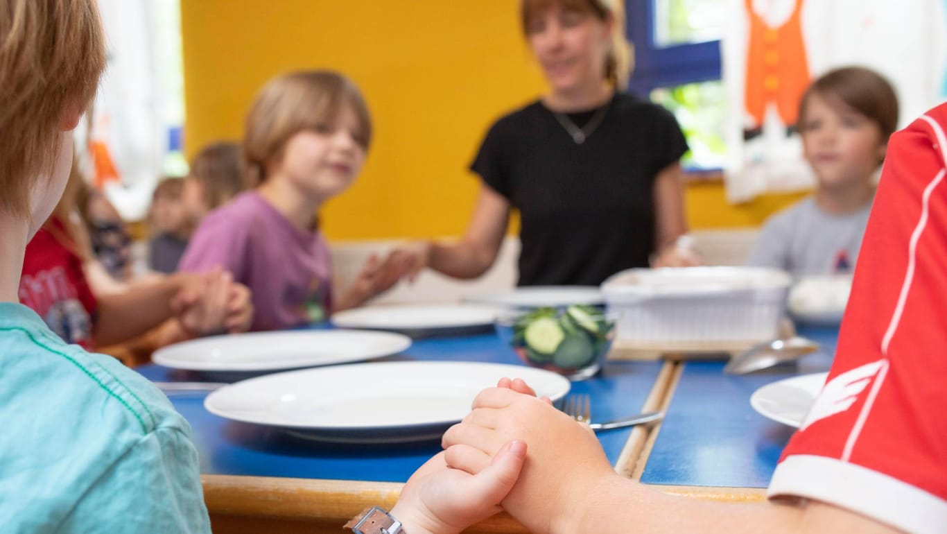 Kinder und Erzieherin im Kindergarten halten sich bei der Hand und sprechen einen Tischspruch: Die Finanzierung kirchlicher Kitas steht auf der Kippe.
