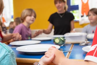 Kinder und Erzieherin im Kindergarten halten sich bei der Hand und sprechen einen Tischspruch: Die Finanzierung kirchlicher Kitas steht auf der Kippe.