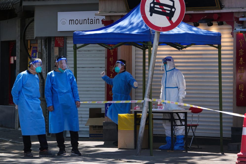 Peking: Helfer in Schutzkleidung stehen vor geschlossenen Einzelhandelsgeschäften an einer Corona-Teststelle.