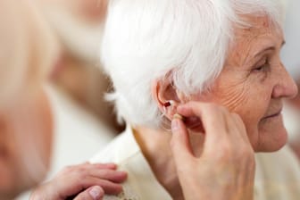 Ärztin setzt Seniorin ein Hörgerät ein: Altersschwerhörigkeit: Im Alter nimmt die Hörleistung ab. Der Prozess beginnt etwa ab dem 50. Lebensjahr. Altersschwerhörigkeit entsteht dadurch, dass die winzig kleinen Haarzellen in der Hörschnecke (Cochlea) nicht mehr vorhanden sind oder beschädigt werden. Ein Hörgerät kann helfen, das Hörvermögen zu optimieren.