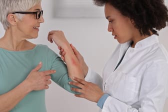 Eine Frau zeigt einer Ärztin eine Hautstelle am Unterarm.