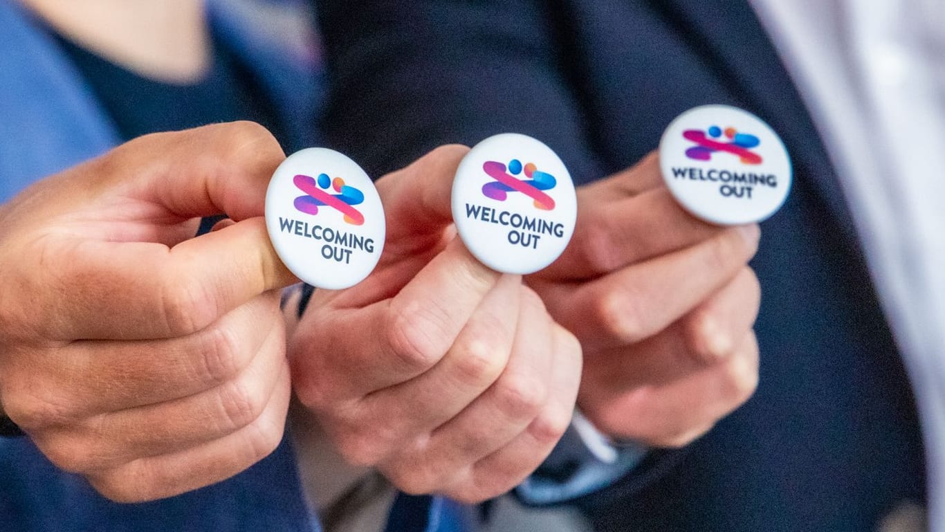 Menschen halten Ansteckbuttons mit dem Logo von "Welcoming out" ins Bild: Das Symbol soll Akzeptanz für queere Menschen sichtbar machen.