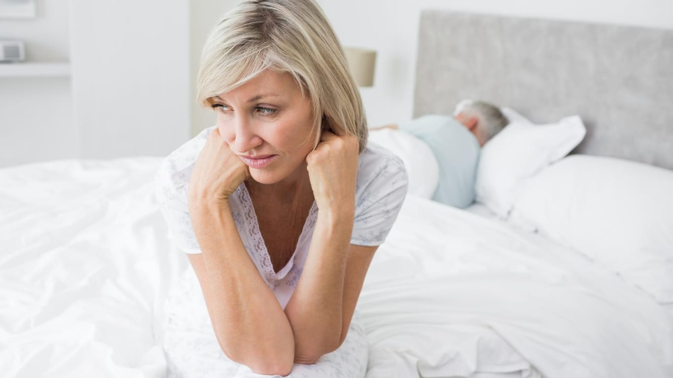 Traurige Frau mittleren Alters auf Bett mit Partner: Scheidentrockenheit kann das Liebesleben belasten. Der Geschlechtsverkehr ist unangenehm und bereitet Frauen oft starke Schmerzen.