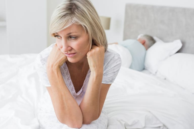 Traurige Frau mittleren Alters auf Bett mit Partner: Scheidentrockenheit kann das Liebesleben belasten. Der Geschlechtsverkehr ist unangenehm und bereitet Frauen oft starke Schmerzen.