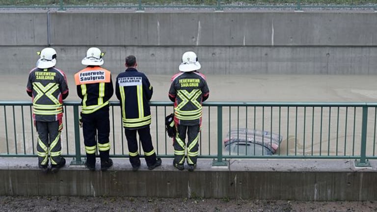 Einsatzkräfte der Feuerwehr schauen auf die Heckklappe eines Autos, das aus dem Wasser auf der teilweise überfluteten Bundesstraße B23 ragt.