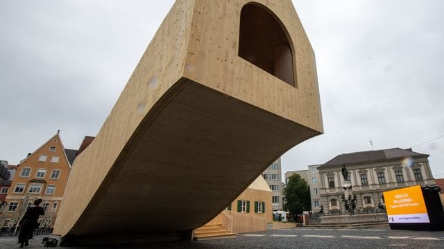 Ein Holzhaus steht freischwebend neben dem Augustusbrunnen.