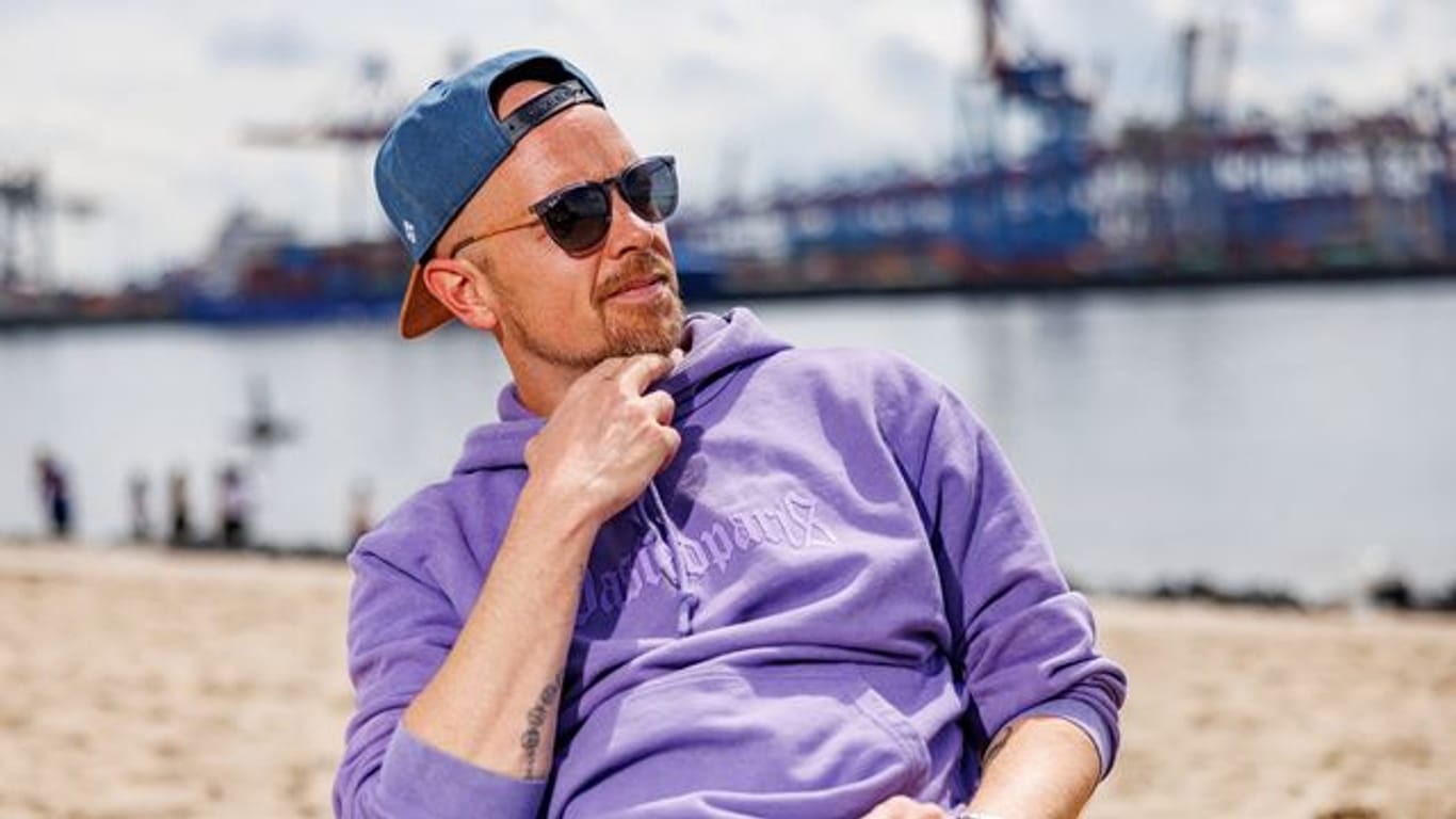 Der Musiker Jan Delay sitzt am Elbstrand in Hamburg vor den Industrieanlagen des Hafens in der Sonne.