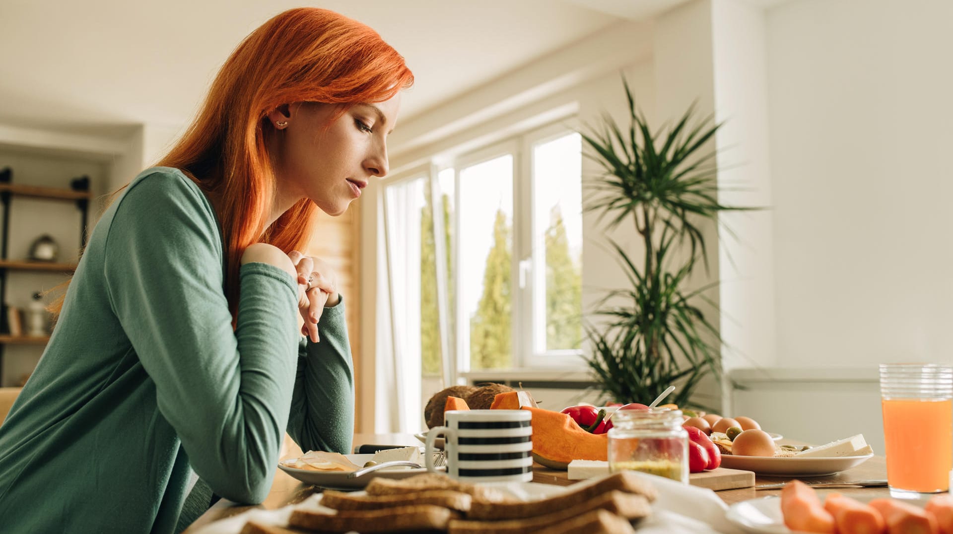 Junge Frau sitzt am Frühstückstisch und schaut nachdenklich auf das Essen.