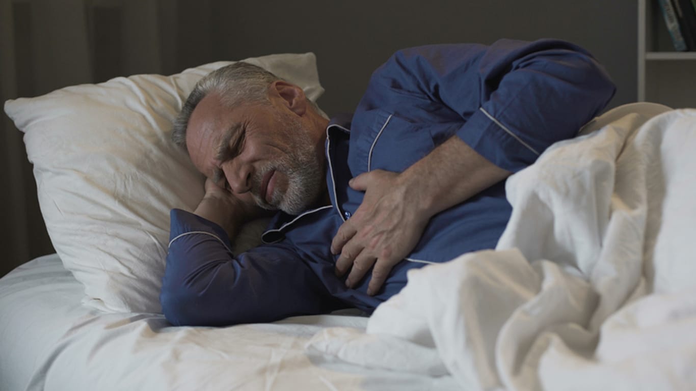 Schlafender Mann greift sich ans Herz. Ausreichend Schlaf ist wichtig, damit der Körper reagiert. Fehlt diese Erholungszeit oder wird sie gestört, drohen Herz-Kreislauf-Erkrankungen.