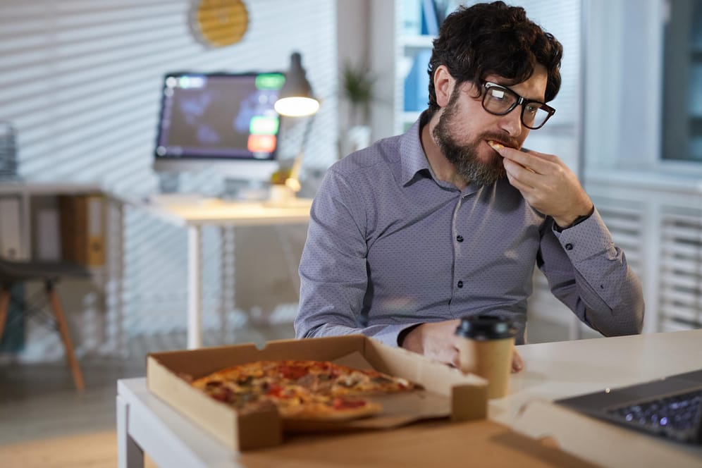 Ein Mann sitzt am Schreibtisch und isst eine Pizza.