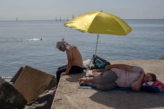 Eine Frau schläft unter einem Schirm auf einem Wellenbrecher vor dem Mittelmeer: Die Menschen suchen Abkühlung.