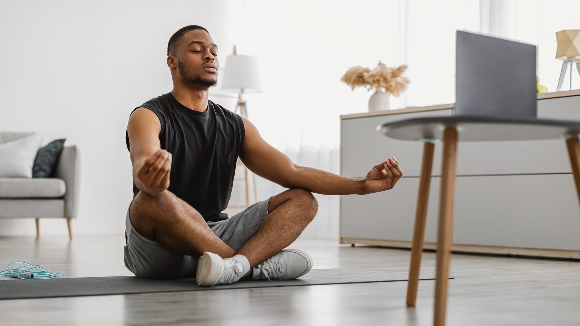 Mann meditiert im Wohnzimmer auf Matte: Meditieren fördert die Konzentration und hilft bei der Stressbewältigung. Dadurch wird die Achtsamkeit im Alltag verbessert.